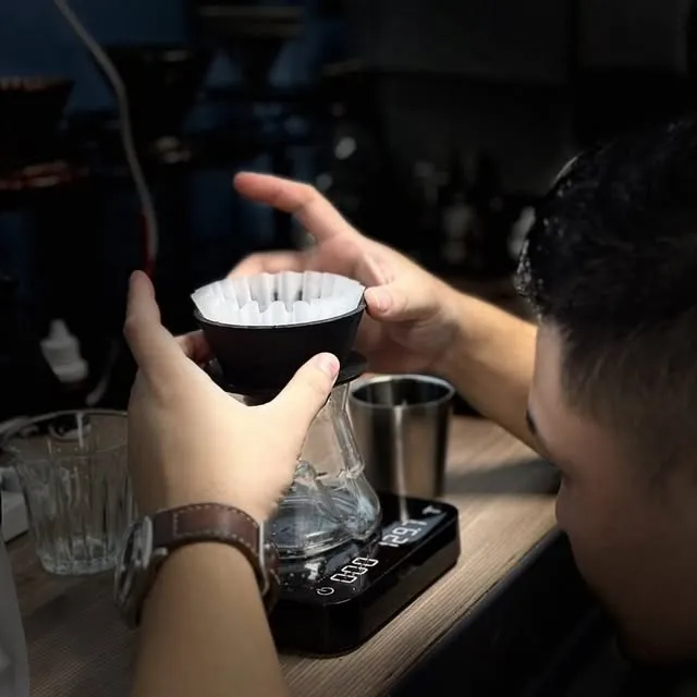 A brewer sets a black ARC Dripper sample, holding a wave filter, on a glass server on a scale