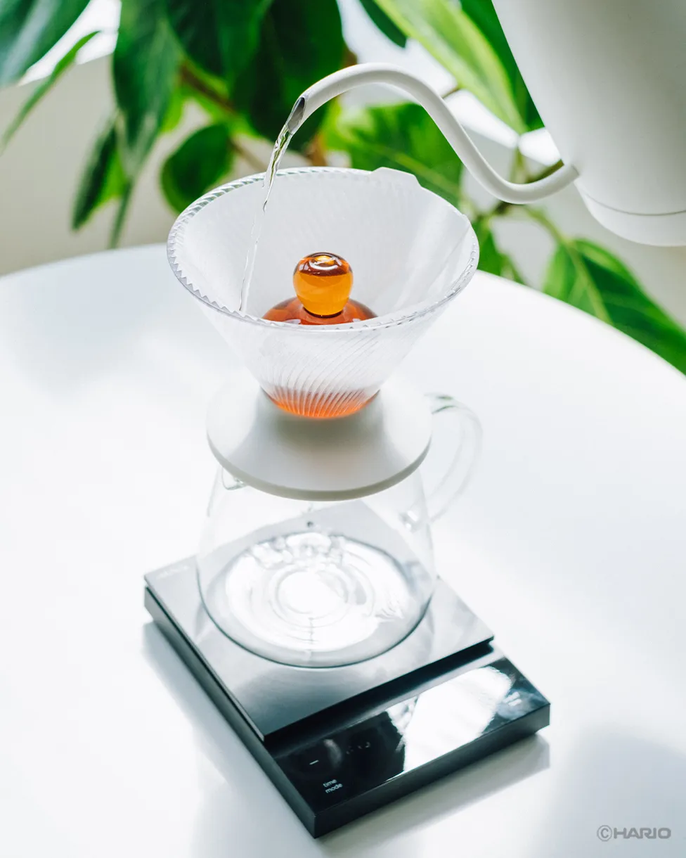 Full setup: white kettle pouring over the amber Rinse Assist in a V60 on a glass server and black scale